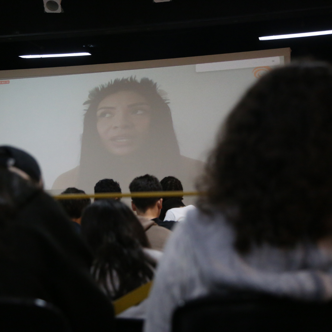 Na foto, estudantes no Teatro Oswald, conversando com a professora indígena Brenda Pataxó, por meio de videochamada exibida em um telão. Fim da descrição.