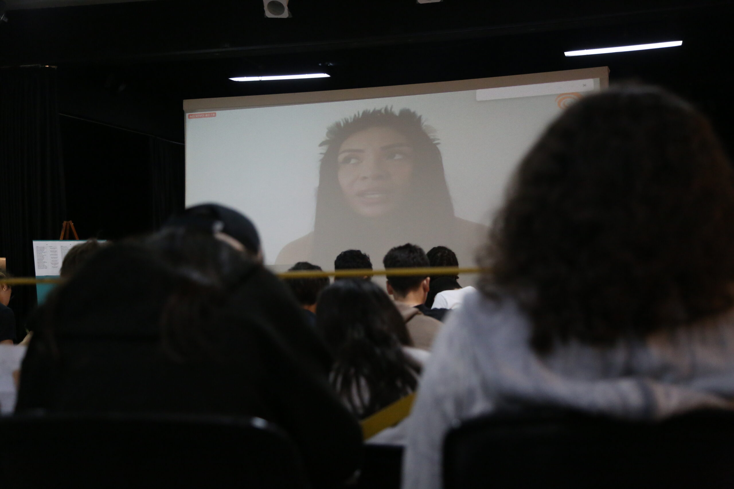 Na foto, estudantes no Teatro Oswald, conversando com a professora indígena Brenda Pataxó, por meio de videochamada exibida em um telão. Fim da descrição.